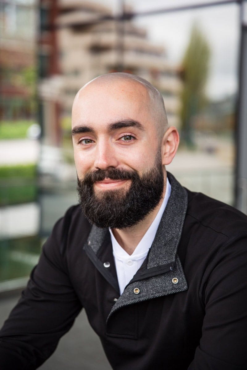 A man with a beard and shaved head, wearing a black jacket and white shirt, smiles at the camera. The background is out of focus, showing windows and greenery.