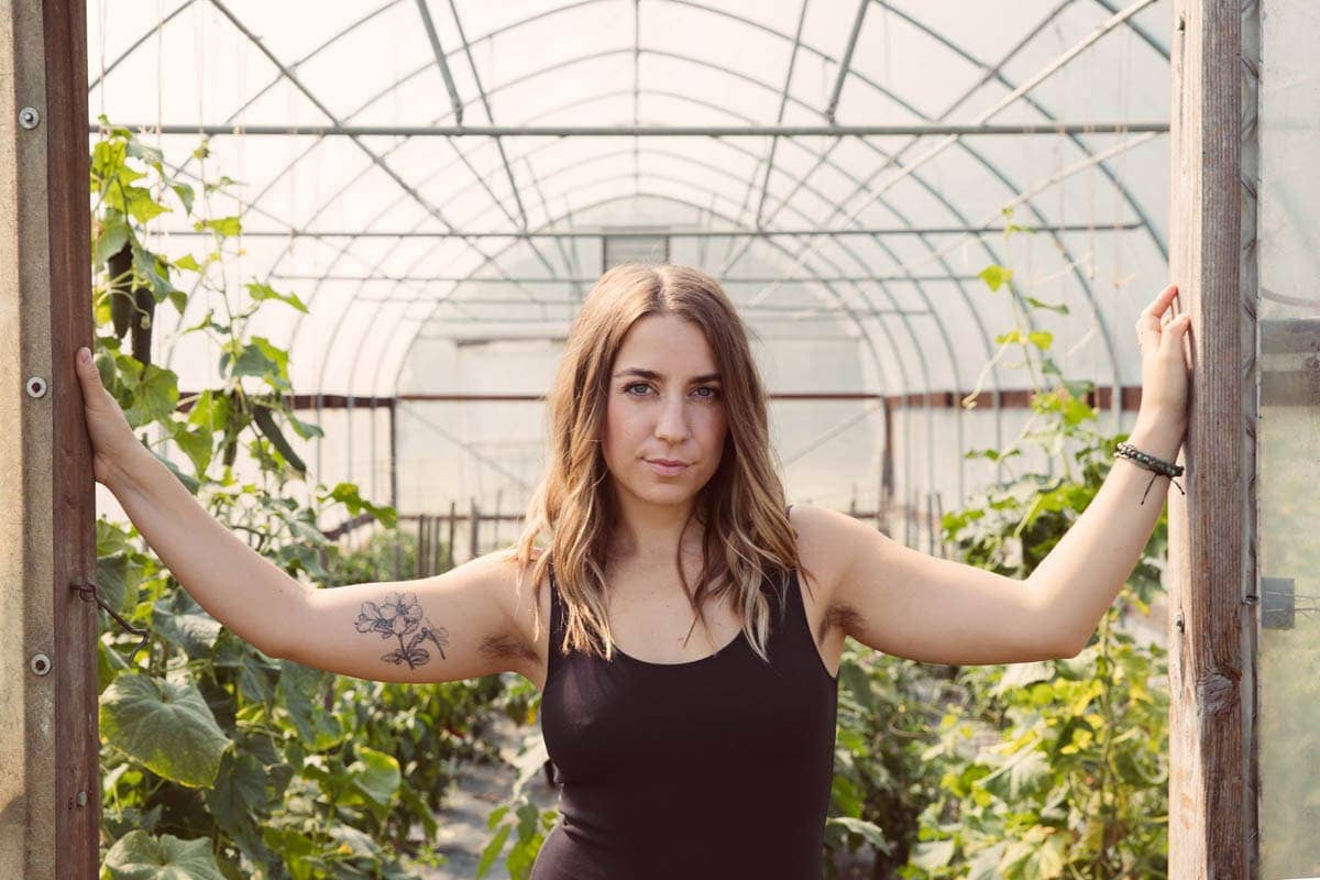 A woman with a floral tattoo on her arm stands in the entrance of a greenhouse, surrounded by green plants, under a curved, transparent roof.