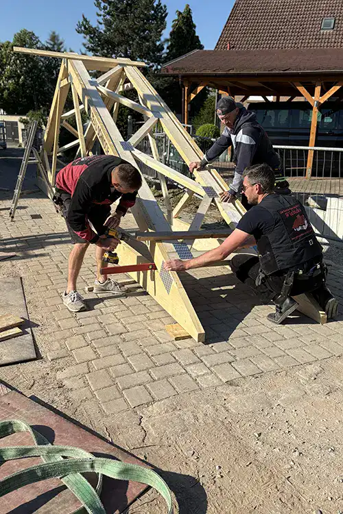 Zwei Personen bauen ein Holzgerüst im Freien auf einem gepflasterten Boden, bei sonnigem Wetter.