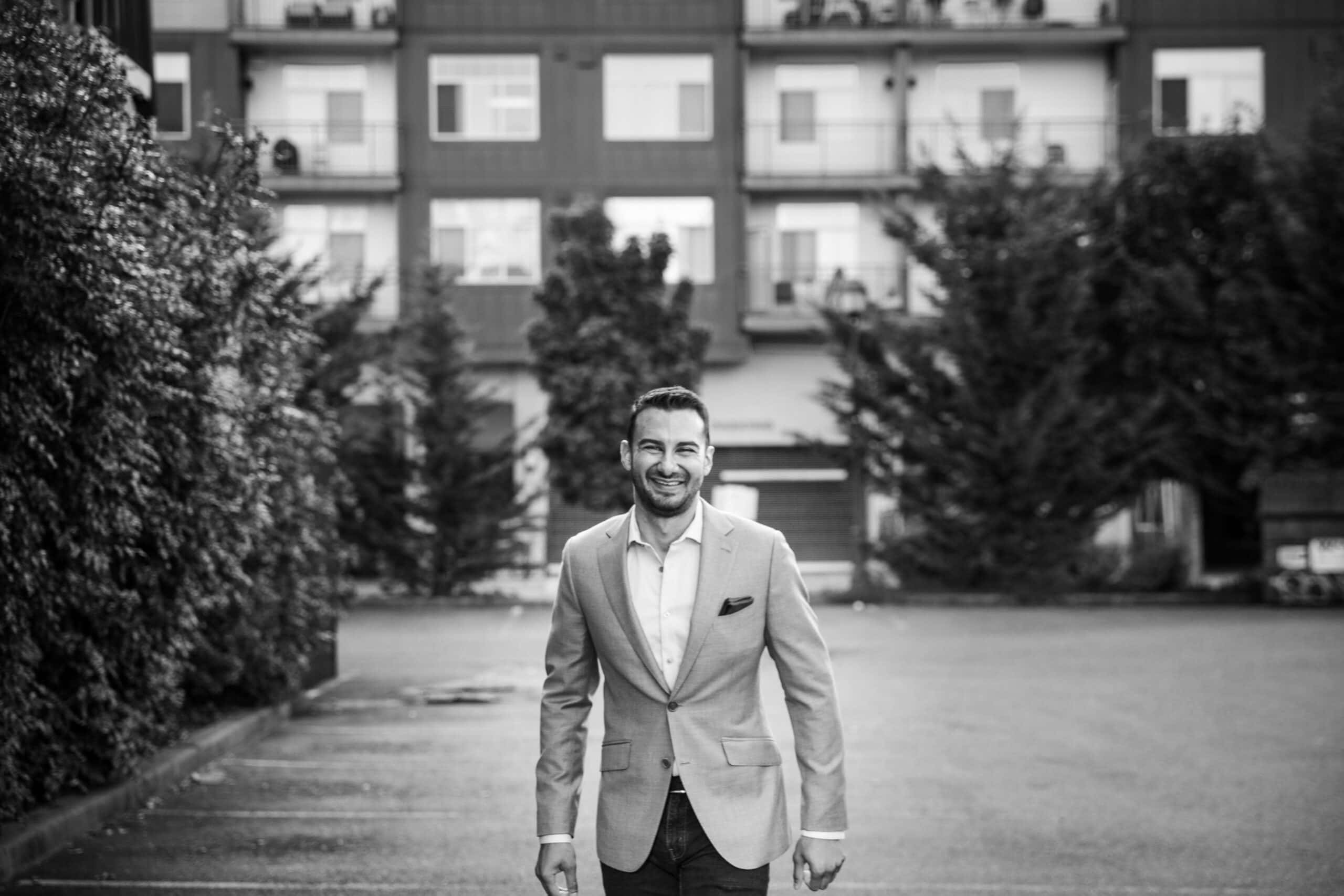 A man wearing a light-colored blazer stands and smiles in an empty parking lot with apartment buildings and trees in the background.