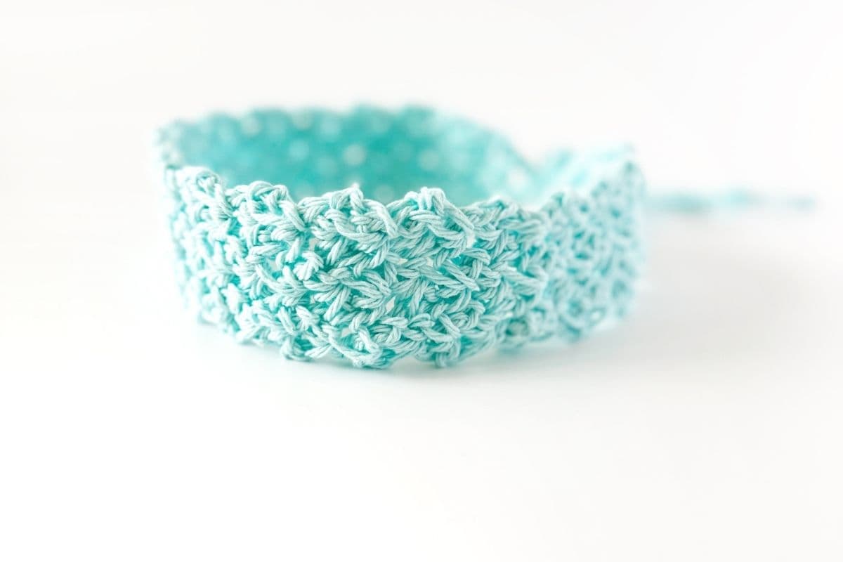 Light blue crocheted headband with a scalloped edge, shown on a white background. The textured pattern and handmade craftsmanship are visible. Free crochet pattern