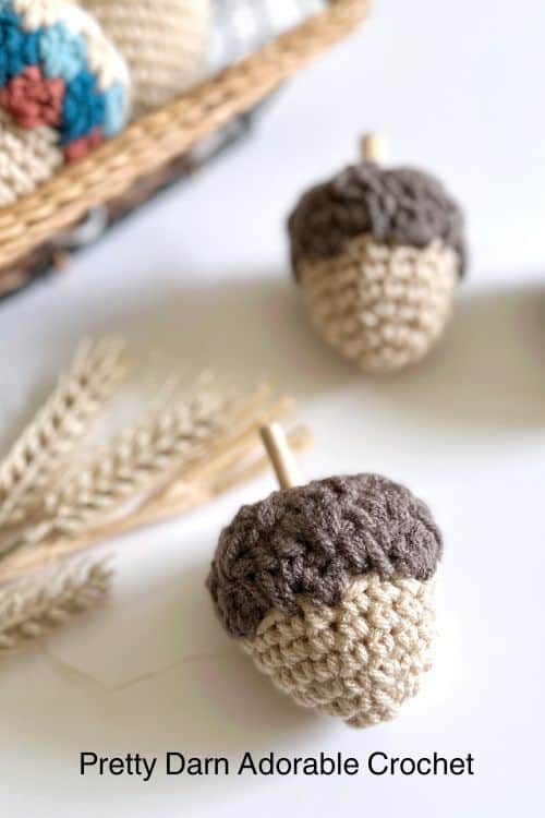 Close-up of two handmade acorns, beige with brown tops, near wheat stalks and a basket. Text: Pretty Darn Adorable Crochet. Free crochet pattern