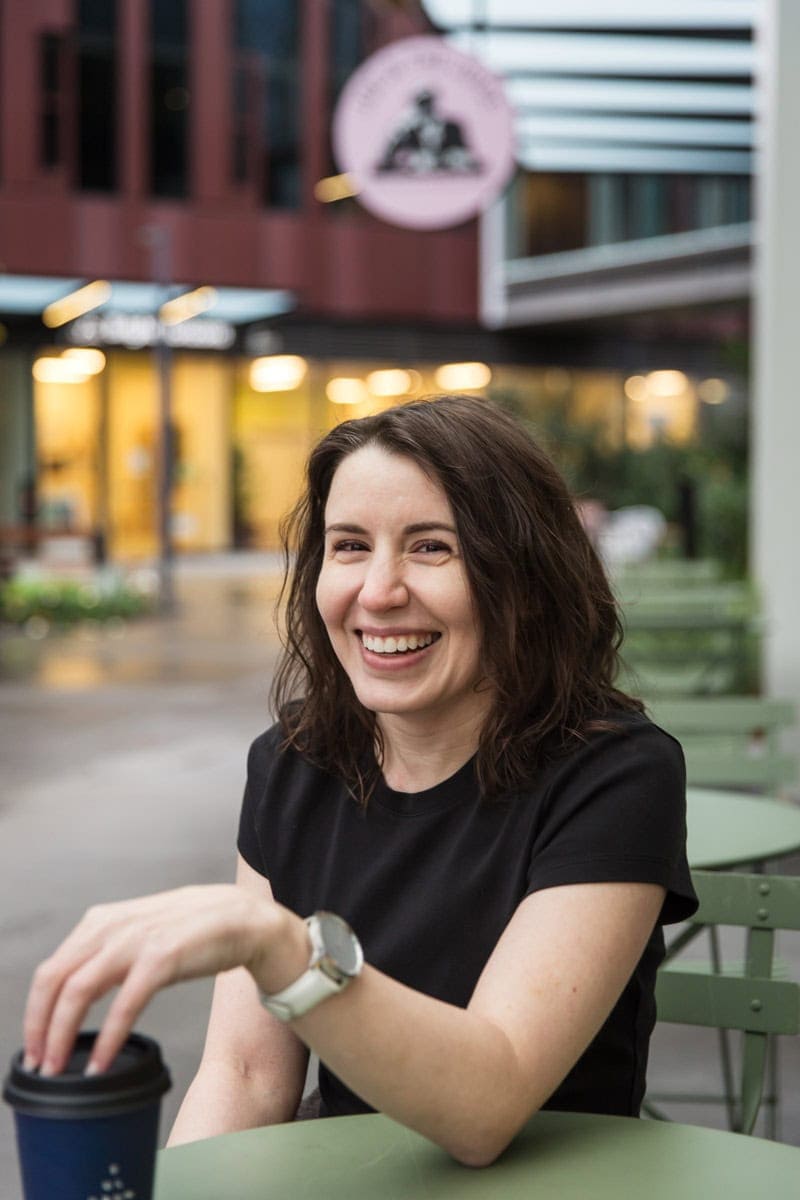 A person with wavy brown hair, wearing a black shirt and silver watch, smiles while sitting at a green table outdoors, holding a coffee cup.