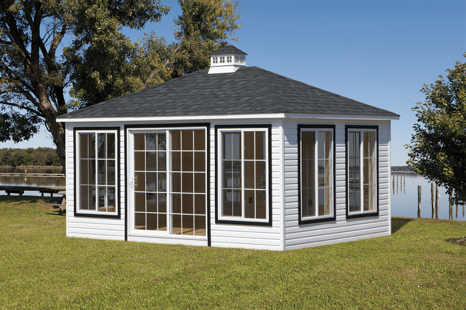 A white, square pavilion with large glass windows and doors stands on a grassy lawn near a lake, with trees in the background.