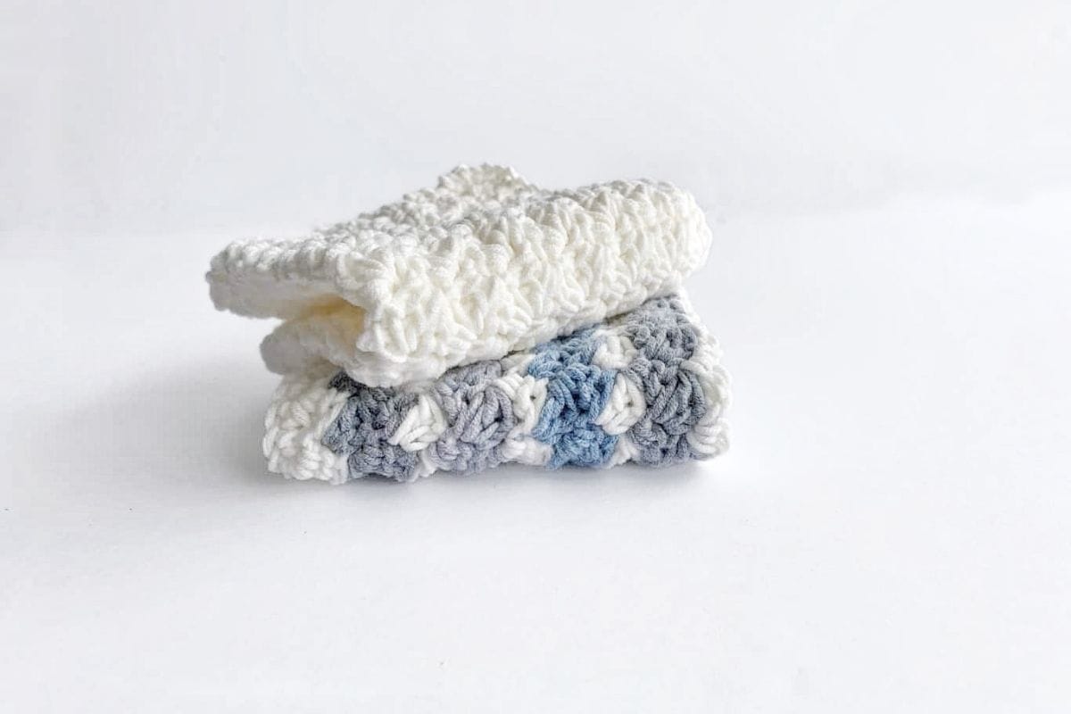 Two folded crocheted washcloths are stacked on a white surface; the top one is off-white, and the bottom has a pattern of white, gray, and blue yarn. No visible text or promotional information appears in the image. Free crochet pattern