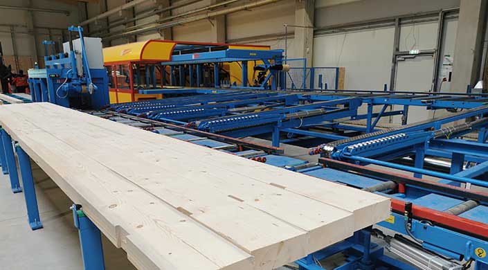 Holzbretter auf einem Förderband in einer Fabrikhalle mit blauer Maschinenanlage im Hintergrund.