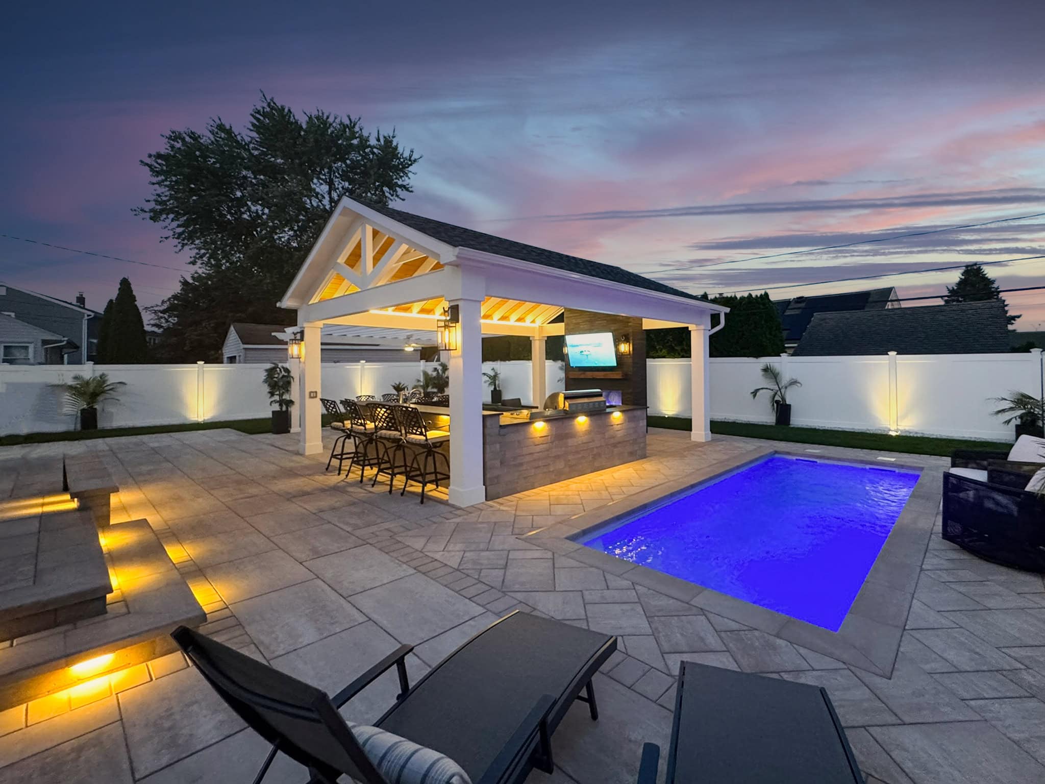 Modern backyard patio with a covered outdoor kitchen, bar seating, illuminated pool, lounge chairs, and soft landscape lighting at dusk.
