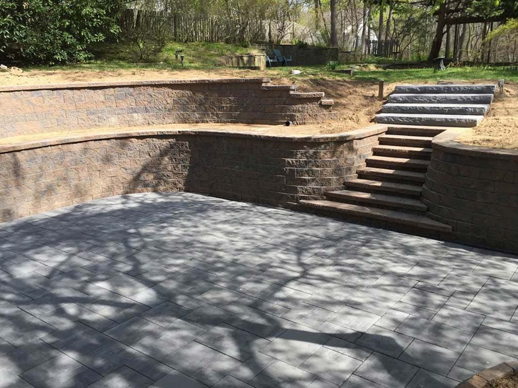 A landscaped backyard features a multi-level stone retaining wall, a paved patio area, and a set of stone steps leading to an upper level in Elwood.