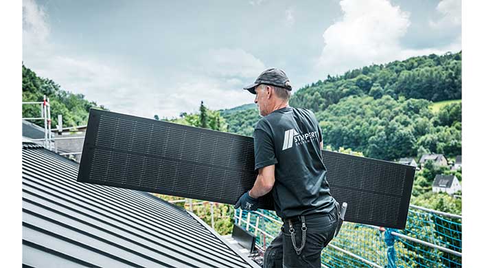 Ein Arbeiter in Arbeitskleidung installiert ein Solarpanel auf einem Dach; im Hintergrund sind Bäume und Hügel sichtbar.