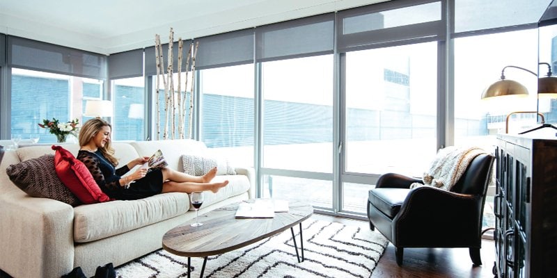 A bright and modern living room with floor-to-ceiling windows that let in natural light. A woman sits comfortably on a cream-colored sofa with red accent pillows, reading a magazine. The room features a sleek coffee table, a stylish black armchair, and contemporary decor. The ambiance exudes relaxation and sophistication, ideal for a leisurely stay or residential living.