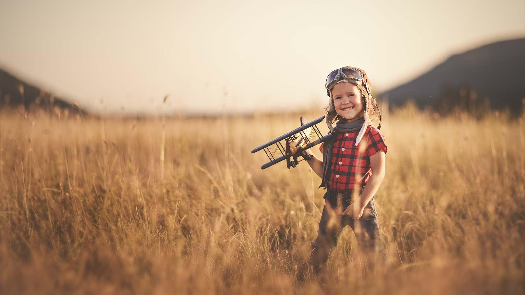 Young child in aviator goggles and scarf stands in a grassy field, holding a toy airplane, with hills and soft sunlight in the background.