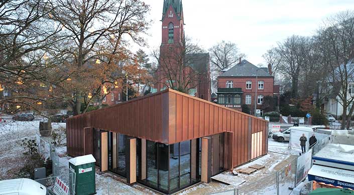 Modernes Gebäude mit Glas- und Kupferfassade im Vordergrund, im Hintergrund eine historische Backsteinkirche mit hohem Turm und spitzem Dach, umgeben von Bäumen.