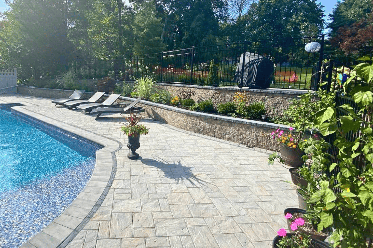 Outdoor pool area with lounge chairs, potted plants, and a stone patio; landscaped garden, retaining walls, and grill visible behind a black metal fence.