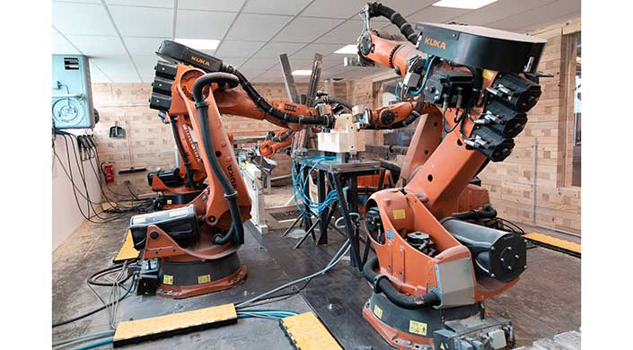 Mehrere orangefarbene Industrieroboter arbeiten in einem modernen Labor mit Tischen, Kabeln und Geräten. Die Umgebung wirkt technisch und auf Automatisierung ausgerichtet.