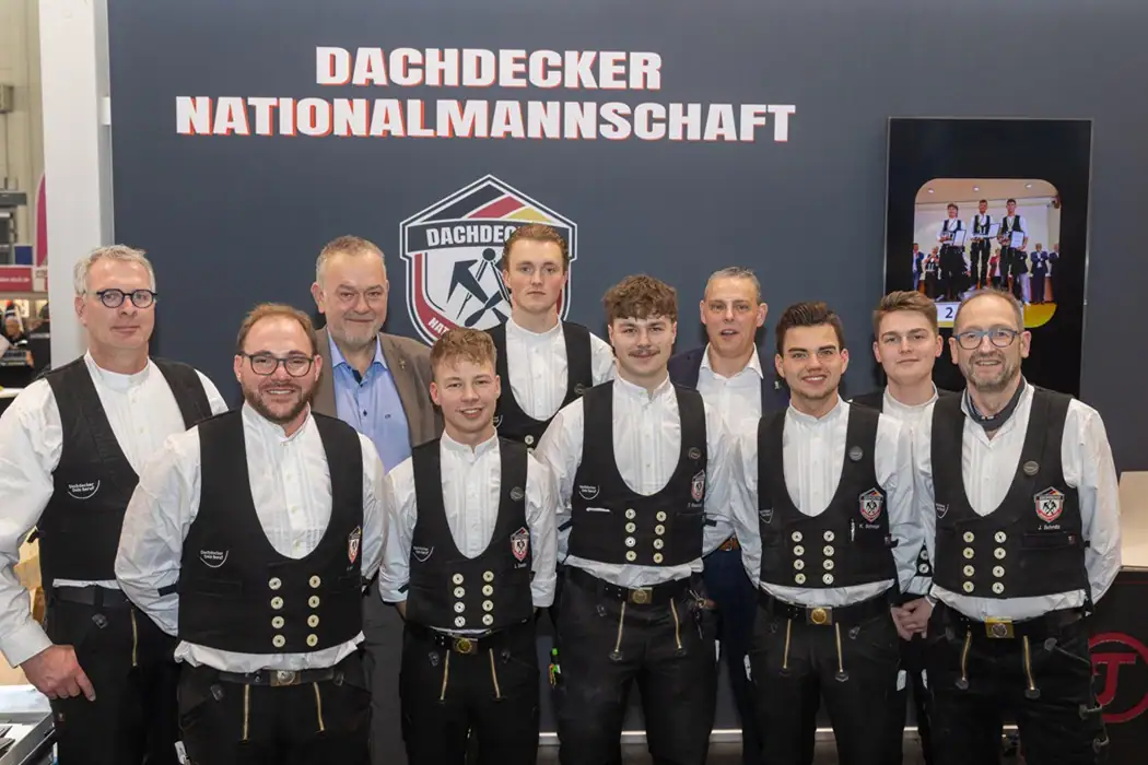 Team und Trainer in traditioneller Arbeitskleidung stehen gemeinsam mit ZVDH-Präsident Dirk Bollwerk im Anzug vor einem Banner mit der Aufschrift „Dachdecker Nationalmannschaft“.