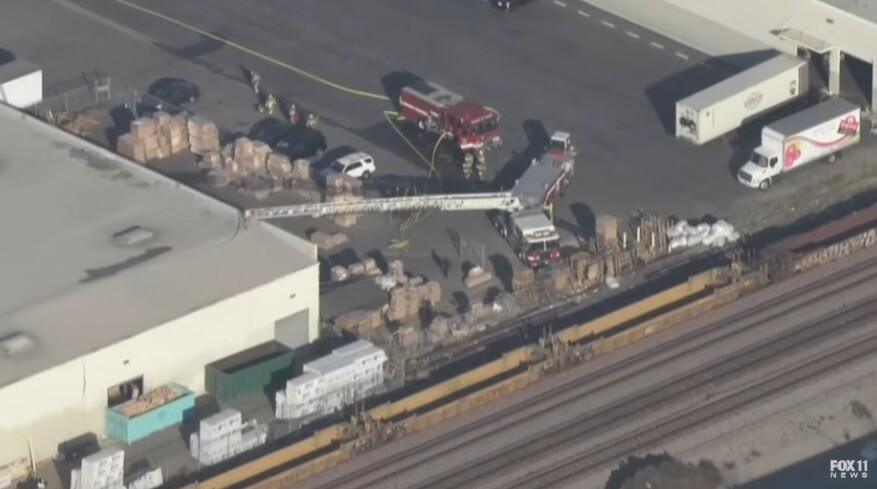 Aerial view of a warehouse fire after a plane crash in Southern California. Fire trucks and emergency personnel are on the scene, with smoke rising from the damaged warehouse roof. A small plane crash in Fullerton. Emergency aid is present.