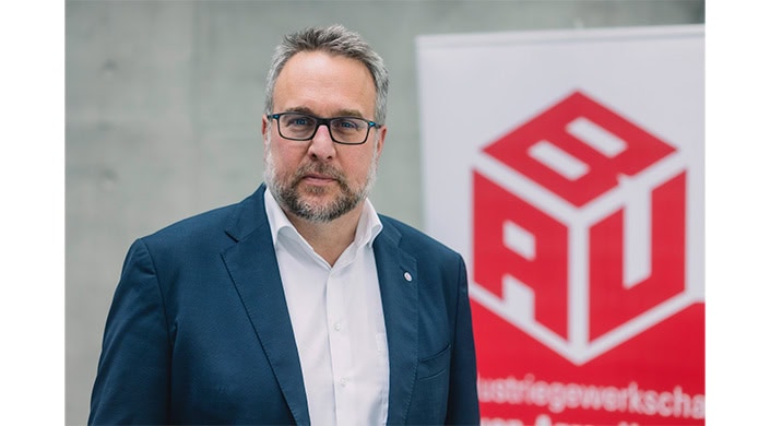 Ein Mann mit Brille, grauem Bart und kurzen Haaren trägt ein weißes Hemd und einen blauen Anzug.