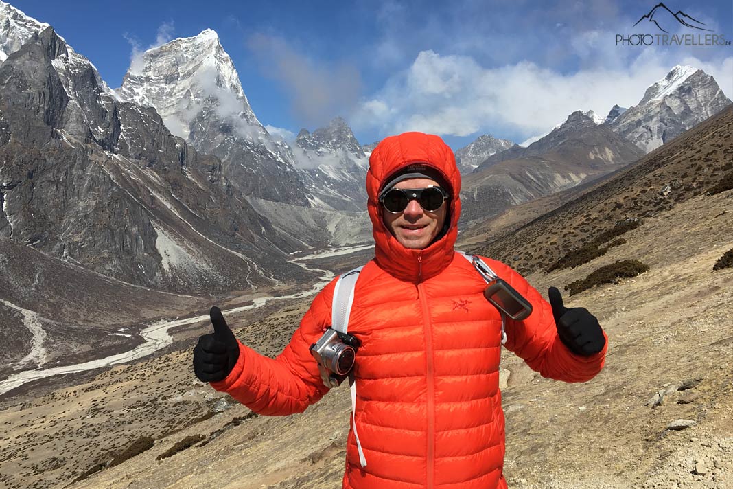 Fotograf Florian Westermann mit oranger Daune und einer Kamera am Rucksack vor imposanten Gipfeln in Nepal