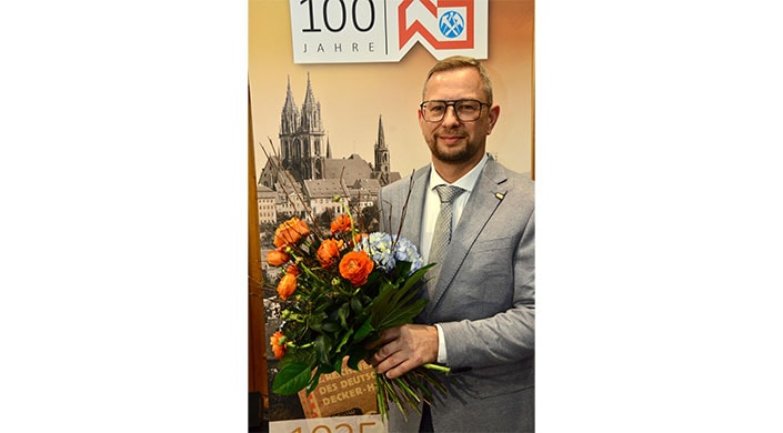 Ein Mann im Anzug hält einen Blumenstrauß, im Hintergrund ein altes Stadtbild und Jubiläumsbanner.