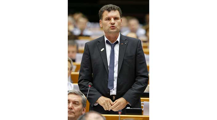 Ein Mann im Anzug spricht stehend in einem Konferenzraum, mit Mikrofonen und anderen Personen im Hintergrund.