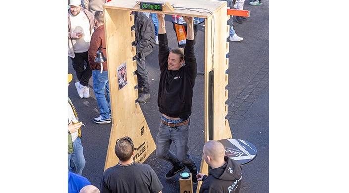 Ein Mann hängt an einer Stange, trägt Jeans und einen schwarzen Hoodie, Menschen stehen um ihn herum, eine Stoppuhr zeigt eine Zeit an.