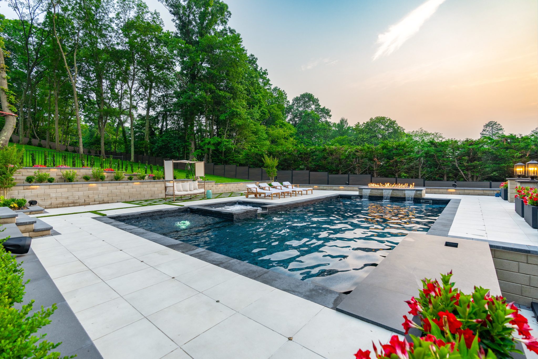 Modern backyard with a rectangular swimming pool, white tile decking, lounge chairs, planters with red flowers, and surrounding trees at sunset.