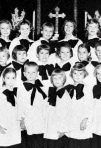 Swedish - ph086 resize Children's choir dressed in white robes with black bows, standing in front of a church altar with a cross, from a historical church photo on the History Museum website.