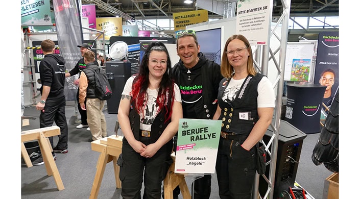 Zwei Menschen stehen lächelnd neben einem Schild mit der Aufschrift „Berufe Rallye - Holzblock ‚nageln‘“ auf einer Messe oder Veranstaltung.