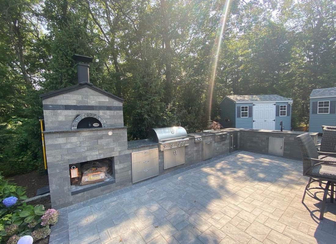 Outdoor kitchen with a pizza oven, grill, and cabinets on a patio surrounded by trees on a sunny day.