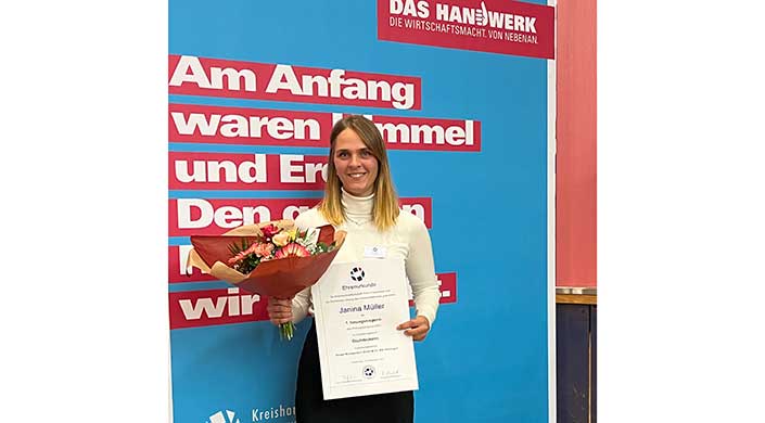 Junge Frau hält eine Urkunde und einen Blumenstrauß vor einer blauen Wand mit rotem Text.