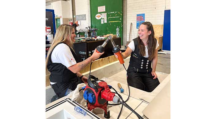 Zwei Personen stoßen mit Werkzeugen in einer Werkstatt an, im Vordergrund befindet sich eine rote Maschine, im Hintergrund ein grünes Fenster und weitere Arbeitsmaterialien.