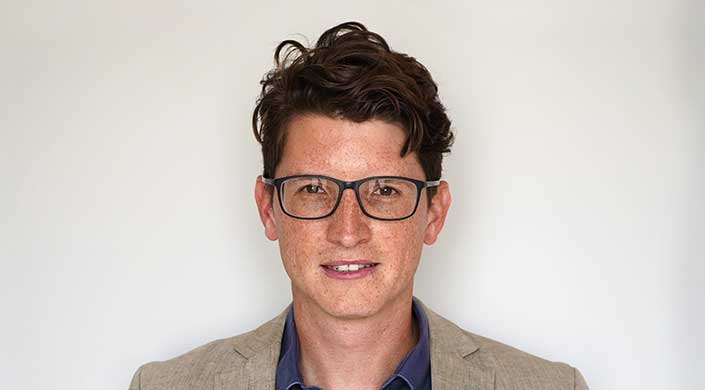 Mann mit braunen Haaren, Brille und Sommersprossen, trägt ein blaues Hemd und schaut direkt in die Kamera.