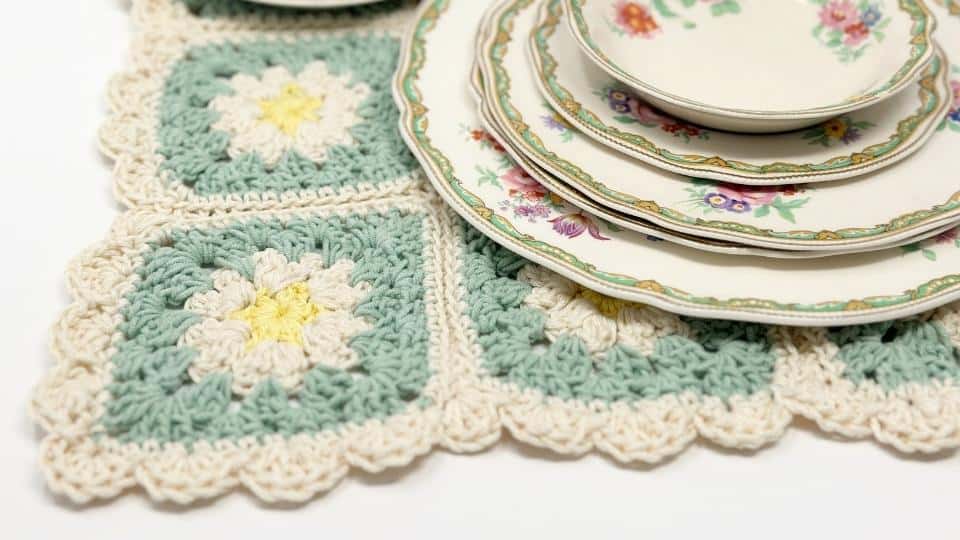 Stacked floral-patterned plates are placed on a pastel-colored crocheted granny square placemat with free patterns, featuring square motifs of white and yellow flowers and a scalloped edge.