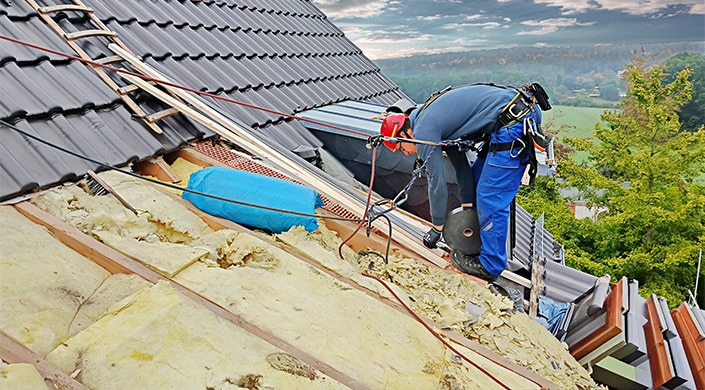 Ein Arbeiter mit Sicherheitsausrüstung arbeitet auf einem Dach mit Isoliermaterial und Dachziegeln bei Tageslicht.