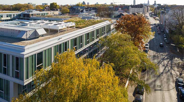 Modernes Gebäude mit begrüntem Dach und großen Fenstern, umgeben von herbstlich gefärbten Bäumen, im Hintergrund eine Stadtlandschaft.