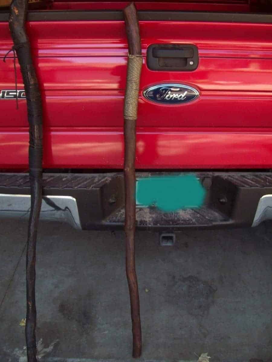 Two handmade walking sticks lean against a red Ford truck. The staff on the left is dark, while the one on the right is wrapped with twine. These oak branch staff items are examples of wooden crafts.