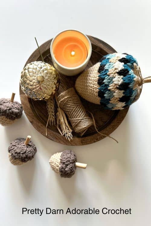 A round wooden tray displays a lit orange candle, gold acorn, twine, and crocheted acorns; text: Pretty Darn Adorable Crochet. Free crochet pattern