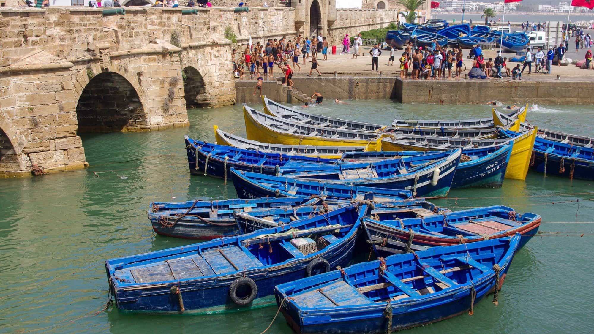 Barcos azules en el puerto de Essaouira, Marruecos, con murallas antiguas y turistas en el fondo.