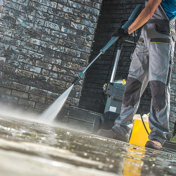 A person using a high-pressure washer to clean a paved surface, with a focus on the water jet and the wet pavement.