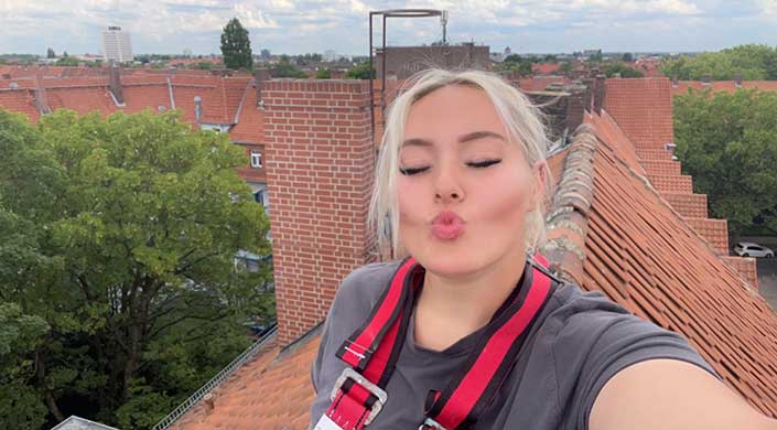 Frau mit blondem Haar macht ein Kussgesicht, trägt ein graues Shirt und ein rotes Sicherheitsgeschirr, im Hintergrund Backsteinmauer und Himmel.