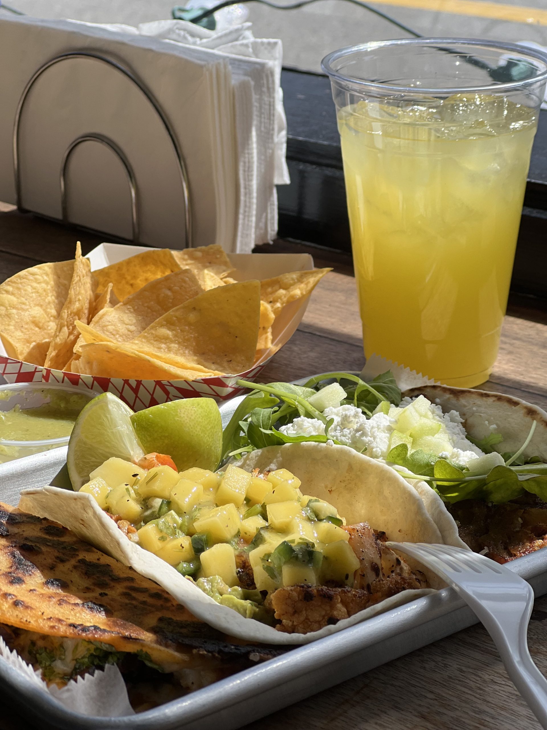 A tray with tacos topped with salsa, chips with salsa dip, and a cup of yellow beverage on a wooden table by a window with napkins in the background. | Mesquite Restaurant Bellport New York