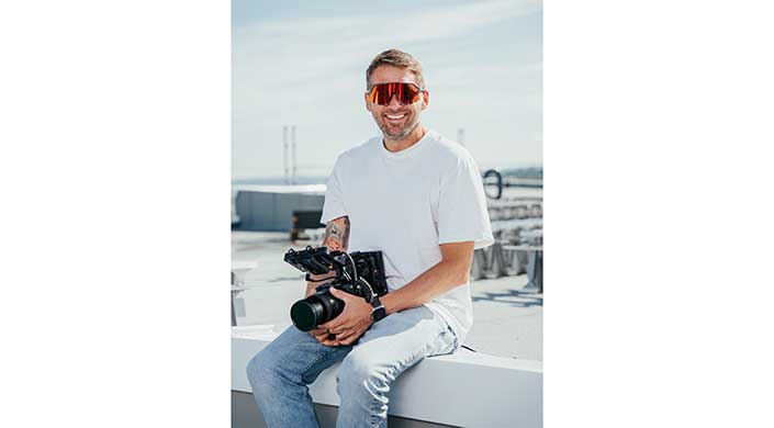 Mann mit Sonnenbrille und weißem T-Shirt hält eine Kamera und sitzt draußen, lächelt in die Kamera.