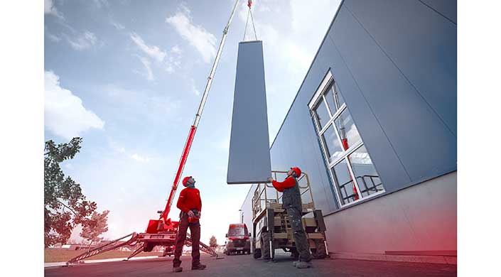 Ein großer Betonfertigteil wird mit einem Kran gehoben, während Arbeiter in Sicherheitskleidung daneben stehen und den Vorgang überwachen.