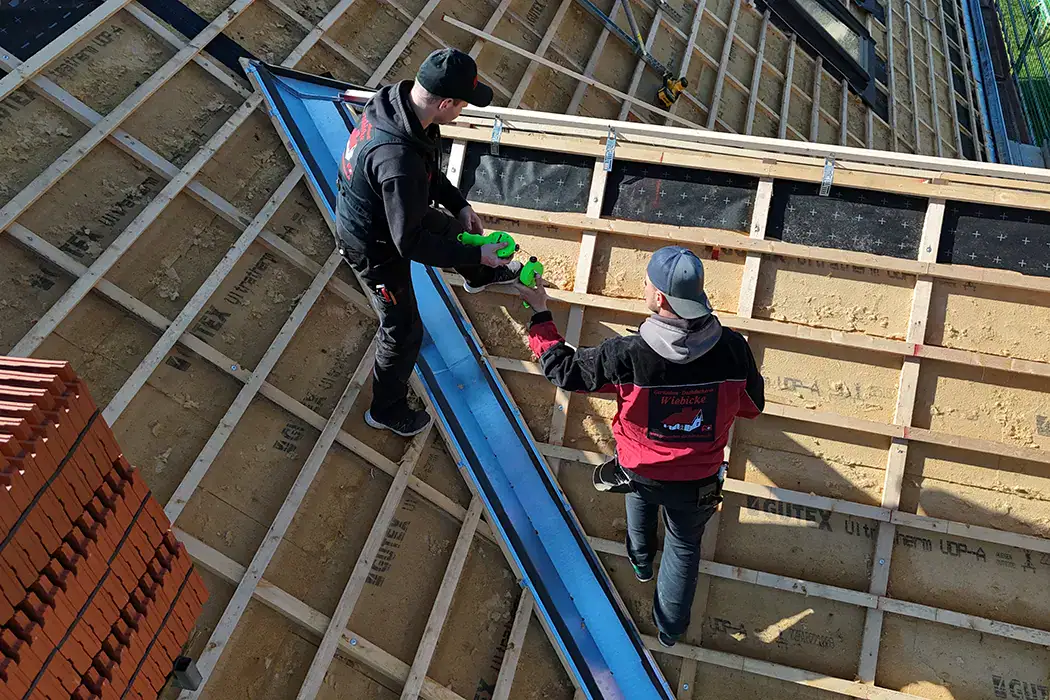 Zwei Personen arbeiten mit grünen Werkzeugen auf einer Baustelle, umrahmt von Holzlatten und blauen Elementen.