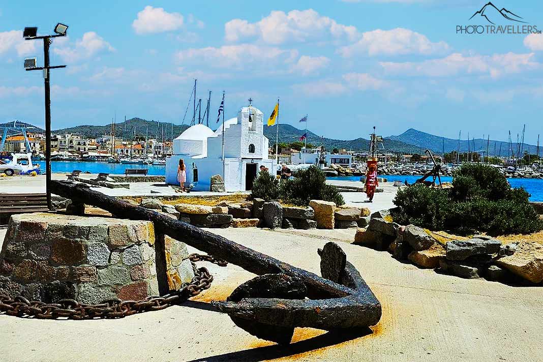 Ein alter Anker liegt zur Deko im Hafen von Ägina mit einer weißen Kirche im Hintergrund