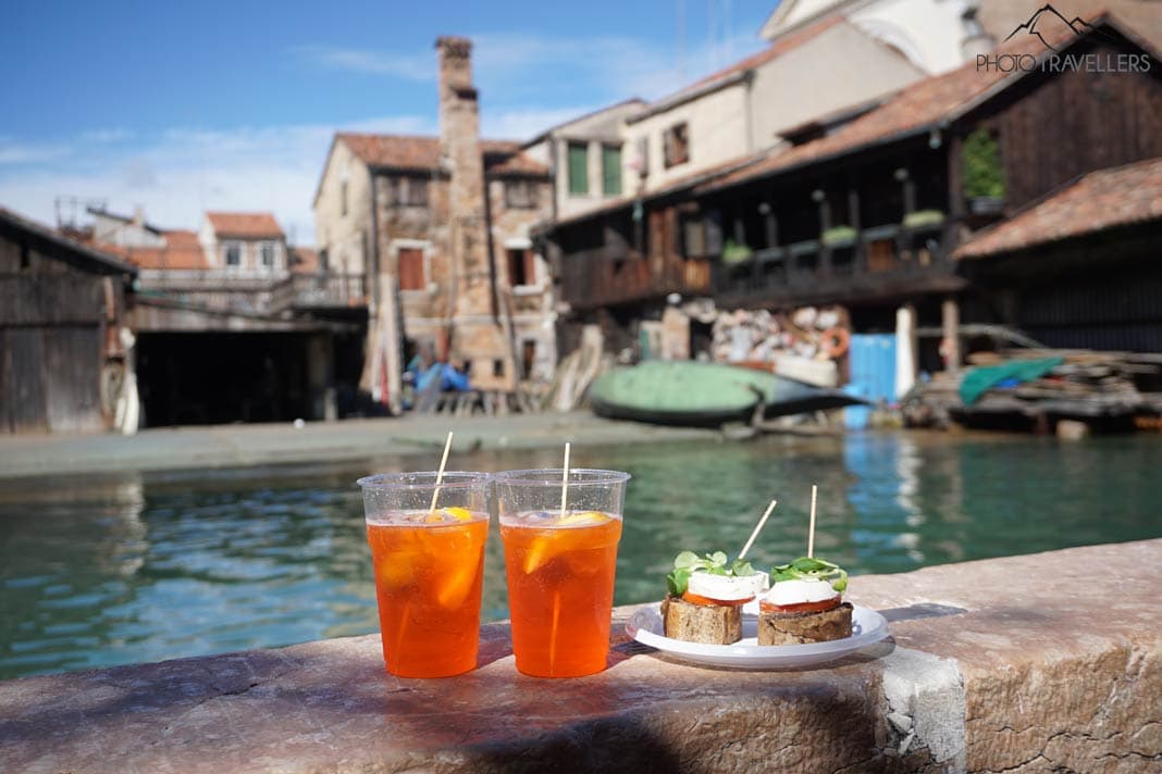 Zwei Aperol Spritz und ein Snack auf einer Mauer, dahinter die alte Gondelwerkstatt Squero di San Trovaso