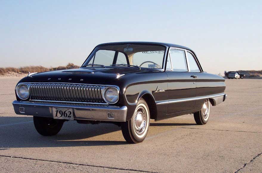 A black vintage 1962 Ford Falcon parked on a paved surface with sand dunes in the background.