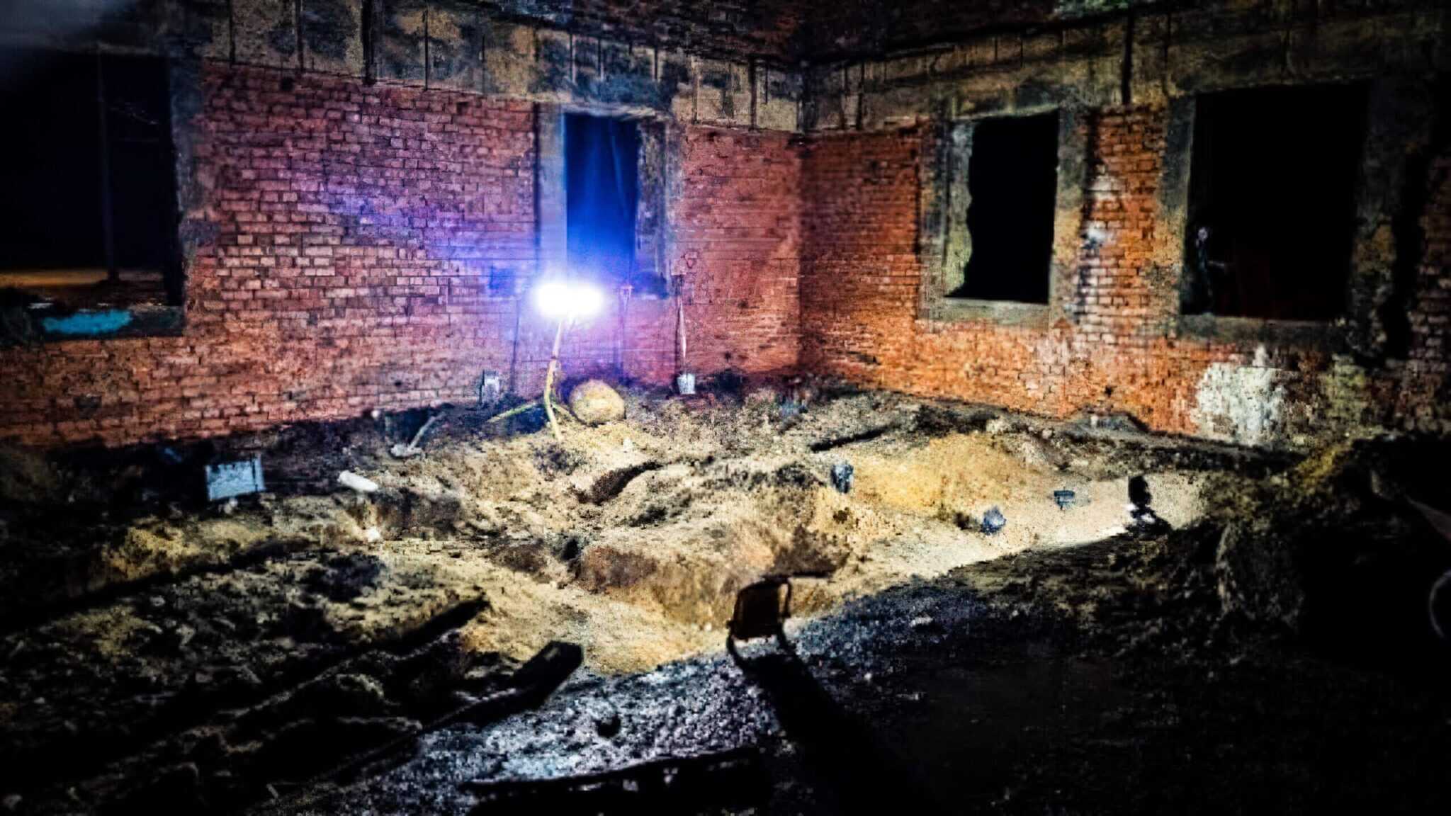 Excavated earth floor lit by a lamp inside a derelict brick building.