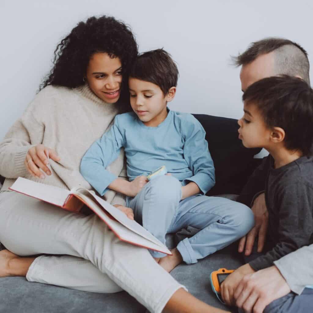 A family sits together, one child in mom's lap and another sitting with dad to look at the book mom is reading.