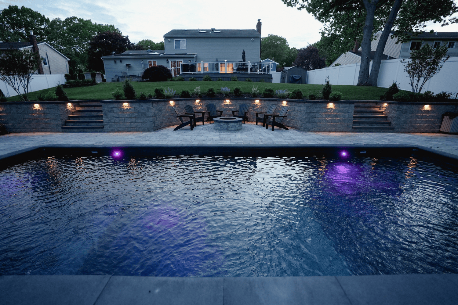 A backyard pool with purple underwater lights, a stone patio bordered by elegant retaining walls, fire pit area with chairs, and a house with a deck in the background at dusk.